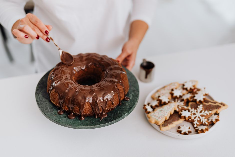 A woman decorates a cake with chocolate glaze beside festive Christmas cookies.
